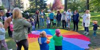 Rodzice i dzieci stoją w ogrodzie przedszkolnym wokół chusty animacyjnej i śpiewają piosenki