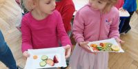 Dzieci wybierają na talerzyki owoce o warzywa, które będą jeść. Na pierwszym planie dwie dziewczynki, które mają pełne talerzyki
