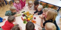 Dzieci i Pani zgromadzone wokół stolika, na środku leża różne owoce i warzywa. Dzieci doświadczają dotykiem owoców i warzy
