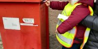 Dziewczynka z pomocą opiekunki wrzuca list do skrzynki pocztowej